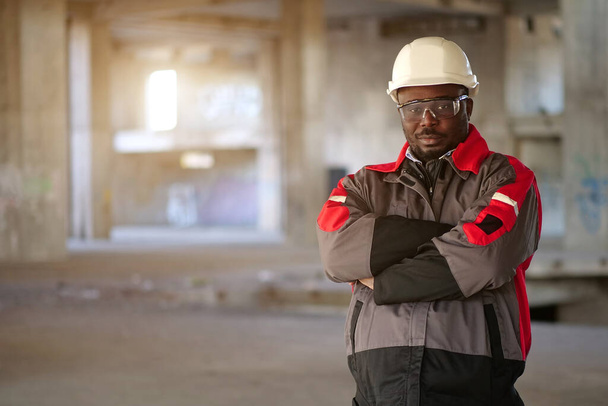Αφροαμερικάνος εργάτης με λευκό σκληρό καπέλο και προστατευτικά γυαλιά στέκεται στο εργοτάξιο και κοιτάζει την κάμερα - Φωτογραφία, εικόνα