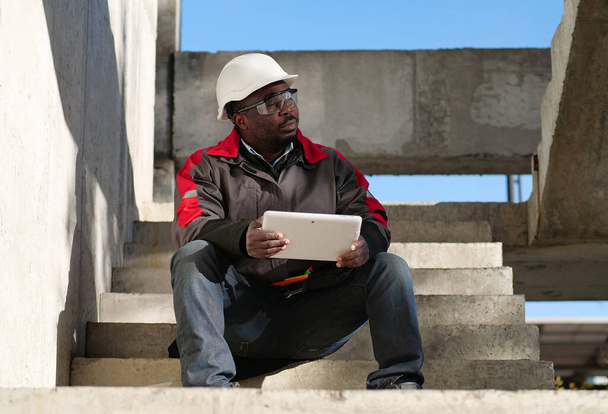 Väsynyt afrikkalainen amerikkalainen työntekijä, jolla on suljetut silmät valkoisessa kypärässä, istuu rakennustyömaalla ja pitää käsissä tabletin PC - Valokuva, kuva