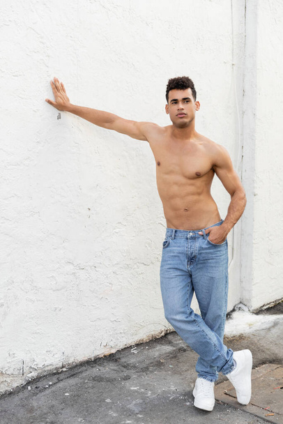 Pełna długość silny i bez koszuli młody Kubańczyk w niebieskich dżinsach pozowanie i dotykanie ściany w Miami  - Zdjęcie, obraz