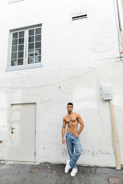 Full length of muscular and shirtless cuban man in blue jeans posing near building in Miami - Photo, Image
