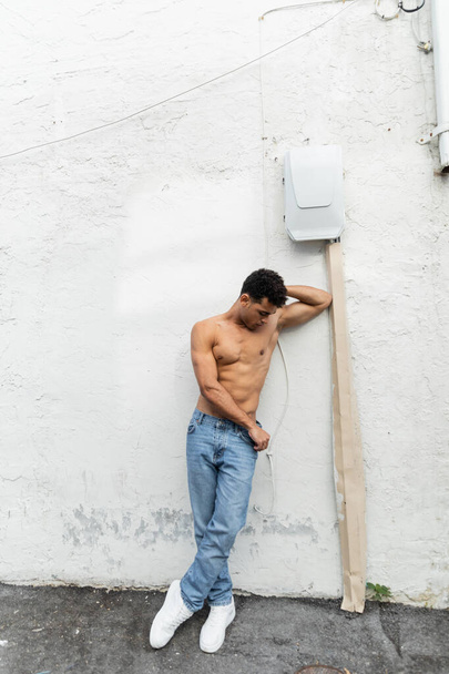 Kubańczyk bez koszuli w dżinsach stojący blisko drutu na białym budynku w Miami latem, umięśniony  - Zdjęcie, obraz