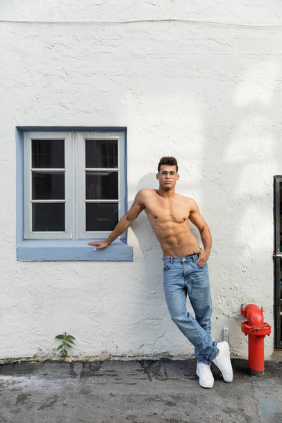 Full length of sexy and shirtless young cuban man in eyeglasses and blue jeans in Miami - Photo, Image