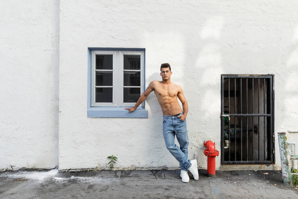 A fehér fal mellett álló félmeztelen, fiatal kubai férfi farmerban és szemüvegben Miamiban. - Fotó, kép