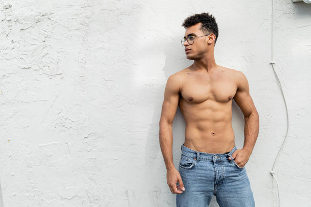 cuban man with athletic body posing in blue jeans and trendy round-shaped eyeglasses near building - Photo, Image