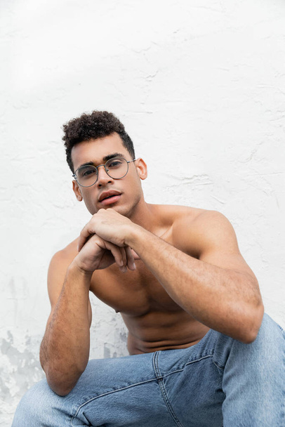 Portrait of relaxed and muscular young cuban man in trendy round-shaped eyeglasses and blue jeans  - Photo, Image