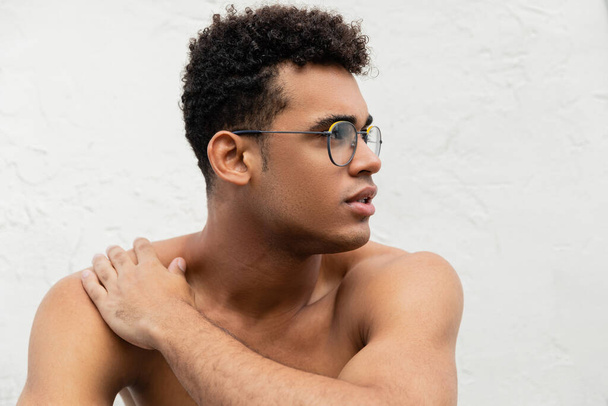 Young shirtless cuban man in trendy round-shaped eyeglasses touching shoulder and looking away - Photo, Image
