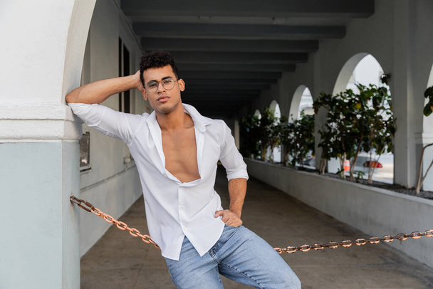 Модный кубинский мужчина в белой рубашке, джинсах и очках держит руку на бедре на городской улице в Майами - Фото, изображение