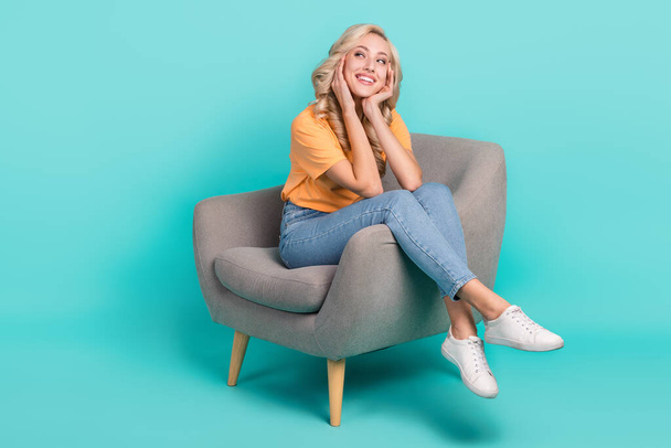 Full length photo of dreamy adorable girl wear orange t-shirt sitting chair arms cheeks looking empty space isolated turquoise color background. - Zdjęcie, obraz