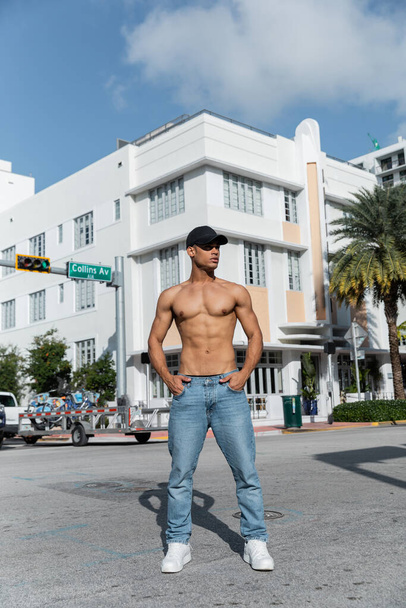 sexy y joven hombre cubano en gorra de béisbol tomados de la mano en bolsillos en la calle urbana en Miami - Foto, imagen
