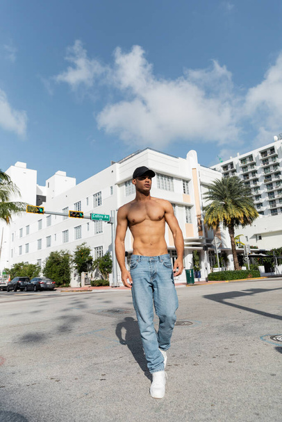 bez koszulki młody Kubańczyk w dżinsach i czapce baseballowej spacerujący po miejskiej ulicy w Miam, lato  - Zdjęcie, obraz