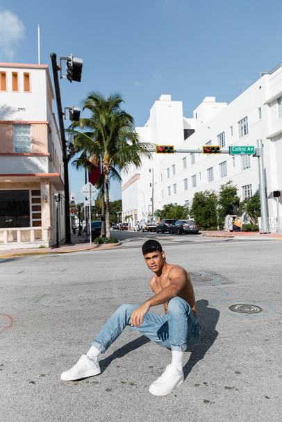 jóképű kubai férfi baseball sapkában és kék farmerban ül az úton Miamiban.  - Fotó, kép