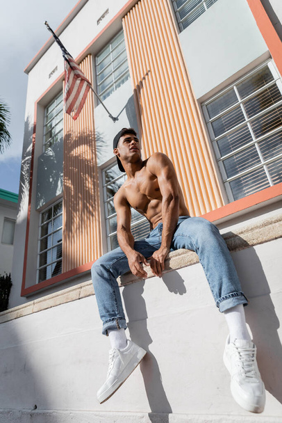kubánský muž v baseballové čepici, džínách a teniskách sedící na parapetu budovy s americkou vlajkou - Fotografie, Obrázek