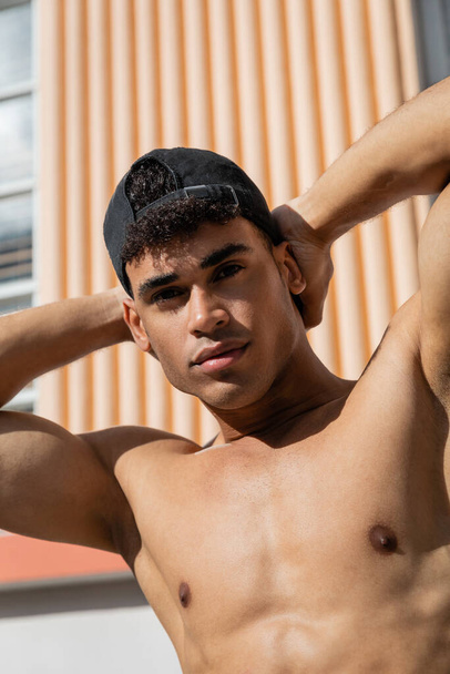 Portret seksownego i umięśnionego młodego kubańczyka w czapce z daszkiem dotykającego szyi i patrzącego w kamerę  - Zdjęcie, obraz