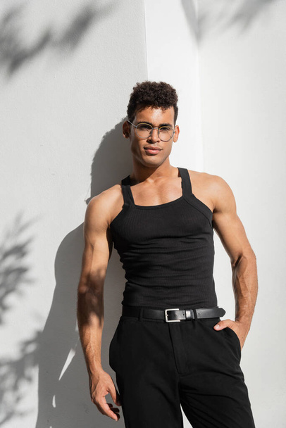 Retrato de homem cubano muscular na moda em óculos e camiseta preta sem mangas posando perto da parede  - Foto, Imagem