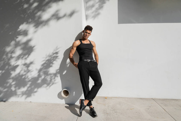 hombre caliente con gafas y camiseta negra sin mangas posando cerca de la pared en Miami, durante el verano - Foto, Imagen