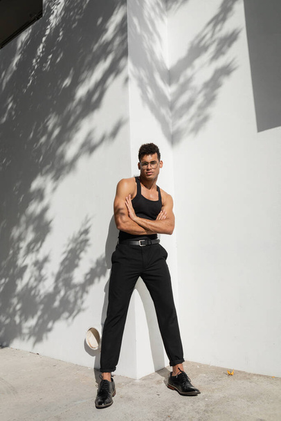 Full length of confident and muscular young cuban man in eyeglasses and trendy outfit crossing arms  - Photo, Image