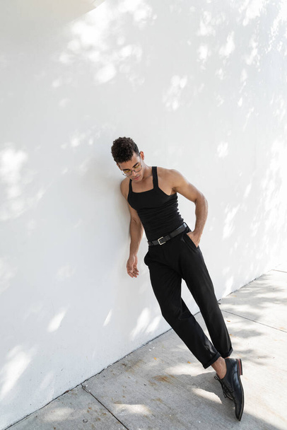 uomo cubano in occhiali da vista e vestito nero che tiene la mano in tasca di pantaloni e in piedi vicino alla parete  - Foto, immagini