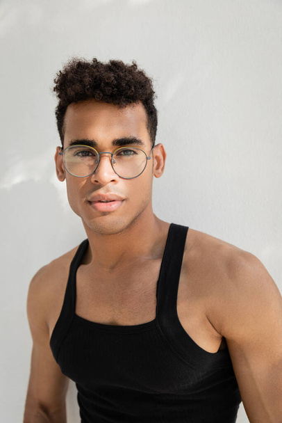 Portret młodego i kręconego kubańczyka w stylowych okularach i koszulce bez rękawów patrzącego w kamerę - Zdjęcie, obraz