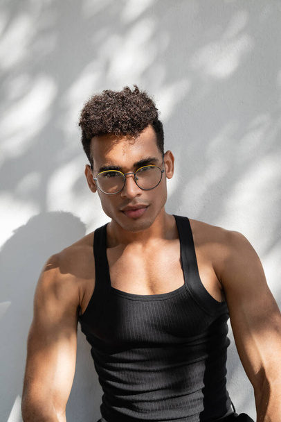 Portrait of cuban man in eyeglasses and trendy black outfit near wall with shadow and sun - Photo, Image
