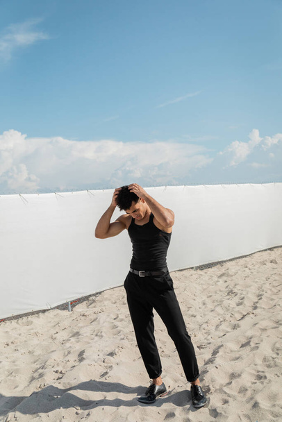 kubanischer Mann im schwarzen Outfit, der am Strand von Miami auf Sand steht und die Haare berührt  - Foto, Bild