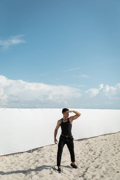Мускулистый и молодой кубинский мужчина в стильной одежде, покрывающий лицо от солнечного света в Майами, южный пляж - Фото, изображение