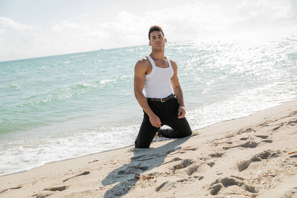 mladý kubánský muž ve stylovém oblečení pózující na pobřeží písku v blízkosti oceánu a v Miami South Beach, Florida - Fotografie, Obrázek