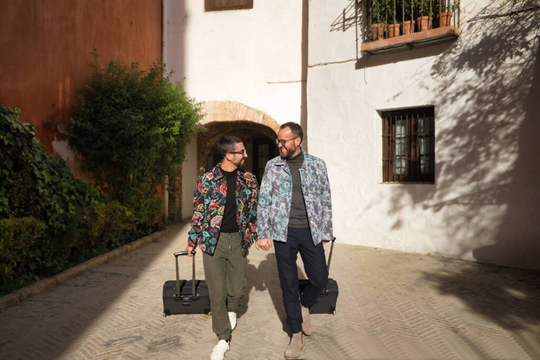 Meleg pár igazi házassága, kéz a kézben sétálnak a városban egy utazási bőrönddel a másik kezükben. Concept lgtb, lgtbiq +, párok, szerelem, utazás. - Fotó, kép