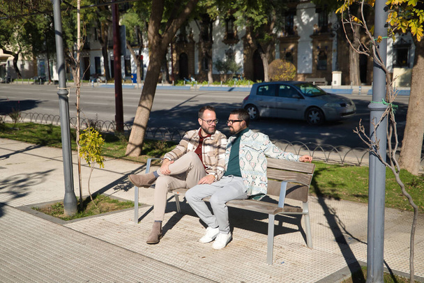 Véritable mariage de couple gay, tenant la main, assis sur un banc en bois, parlant complices et heureux. Concept lgtb, lgtbiq +, couples, amoureux, complicité. - Photo, image