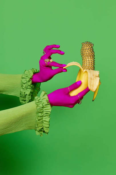 Portrait of colored female hands wearing rings holding gold banana over green studio background. Trash pop art, bright colors. Concept of meal, jewelry, fruits, diet, fashion, beauty, sales, ad - Photo, Image