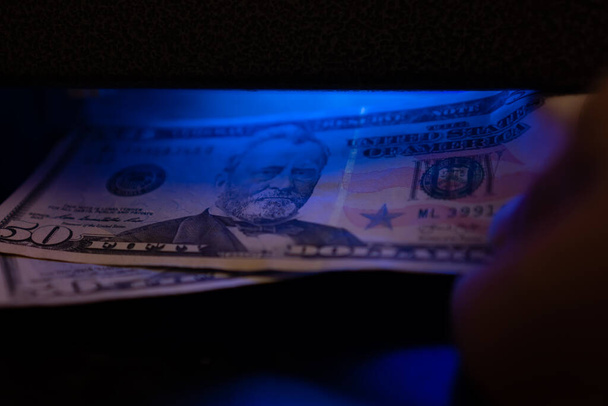 Een close-up van dollars onder een ultraviolette lamp. Inspectie van een dollarbiljet met behulp van een gelddetector. - Foto, afbeelding