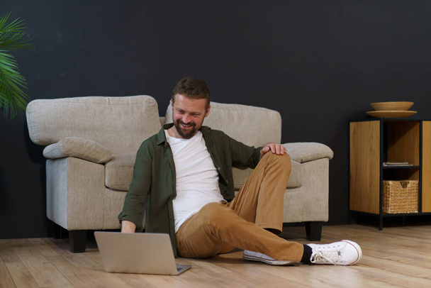 Hombre sentado en el piso cerca del sofá en casa y trabajando en el ordenador portátil. Es freelancer o trabajador remoto, utilizando la tecnología e internet para hacer trabajo. Foto de alta calidad - Foto, imagen