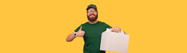 Banner amarelo estúdio tiro de um homem bonito segurando uma caixa de bolo de entrega e polegar para cima. - Foto, Imagem