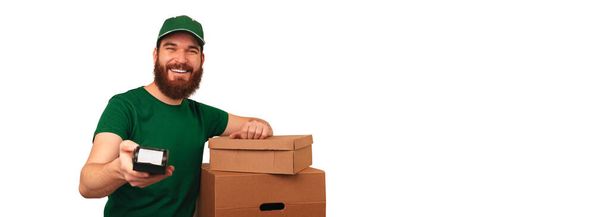 Handsome young delivery man is handing you POS terminal while leaning on boxes. Banner size shot over white background. - Foto, immagini