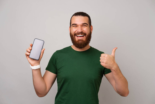 Ευρεία χαμογελαστός άνθρωπος φορώντας πράσινο μπλουζάκι δείχνει κενή οθόνη του τηλεφώνου και τον αντίχειρα προς τα πάνω. - Φωτογραφία, εικόνα