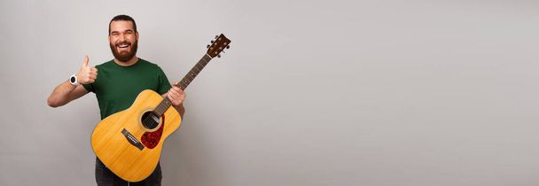 Banner grabado con espacio de copia de un hombre barbudo sostiene una guitarra acústica y un pulgar hacia arriba. - Foto, imagen