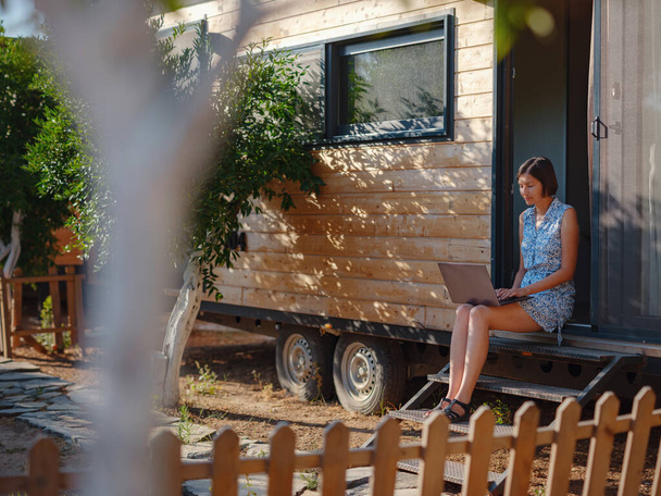 Happy young asian woman with laptop resting outdoors near tiny house, weekend away and remote office idea. Tiny houses and Small Living concept. summer garden - Photo, Image