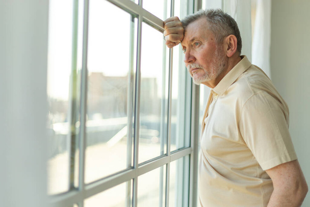 Sad tired lonely disappointed older senior man at home alone. Unhappy grandfather looking sadly outside window experiencing grief relative death bad news. Stressed elder man suffering from loneliness - Foto, Imagen
