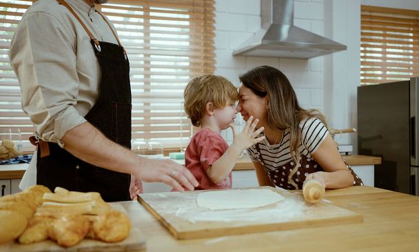 Πατέρας και μητέρα διδάσκουν τον μικρό γιο ζυμώνοντας ζύμη στον πάγκο της κουζίνας στο σπίτι. Γονείς και παιδί αγόρι απολαμβάνουν και διασκεδάζουν εσωτερική δραστηριότητα μαγείρεμα μαζί. - Φωτογραφία, εικόνα