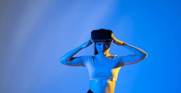 Young woman wearing virtual reality goggles and speasing with a friends in the metaverse. Famale in a black top exploring virtual reality video games in a studio - Фото, изображение