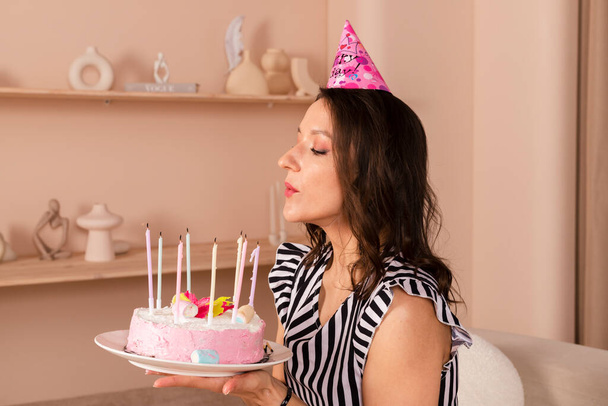 Hermosa mujer morena celebrando su cumpleaños, vestida con un vestido corto a rayas y una gorra de cumpleaños, sostiene la torta rosa y pidiendo un deseo, sopla las velas. - Foto, imagen