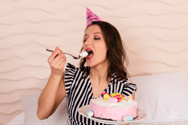 Όμορφη μελαχρινή γυναίκα γιορτάζει τα γενέθλιά της, ντυμένη με ένα ριγέ κοντό φόρεμα και ένα καπέλο γενεθλίων, κάθεται στο κρεβάτι και τρώει ένα κομμάτι από το ροζ κέικ - Φωτογραφία, εικόνα
