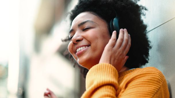 Чорна жінка з навушниками для прослуховування музики в місті для подорожей, мотивації та щасливого мислення. Молода людина на міській вулиці з фоном будівель під час передачі подкасту або аудіо на відкритому повітрі
. - Фото, зображення