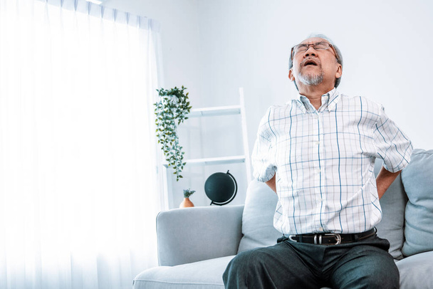 An agonizing senior man in need of assistance while sitting on his sofa at home, suffering from back pain. Senior care, nursing home for pensioners, deteriorating health of old age. - 写真・画像