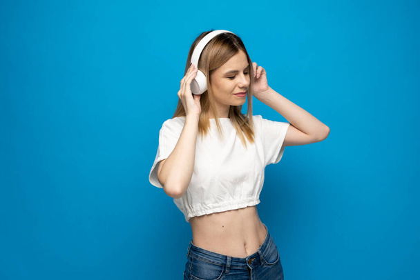 Young blonde woman with headphones listening music .Music teenager girl dancing against isolated blue background - Foto, imagen