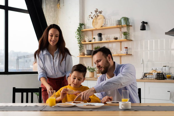 A little boy eats sandwiches for breakfast while sitting at the kitchen table with his parents. A man explains something to his son during breakfast. High quality photo - Photo, Image