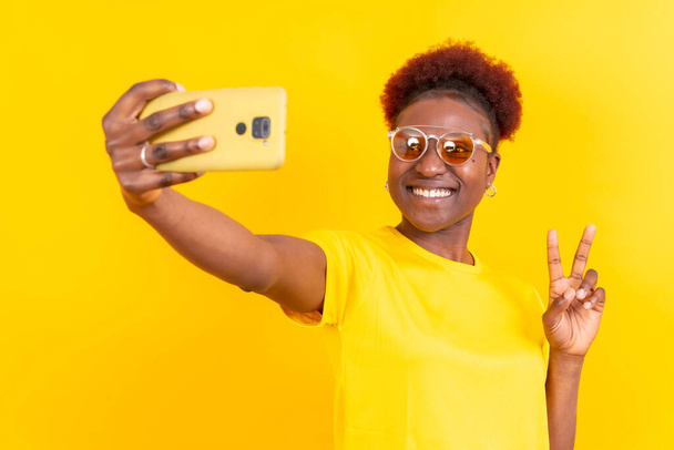 Young african american woman isolated on a yellow background smiling and making a selfie, studio shoot - Fotoğraf, Görsel