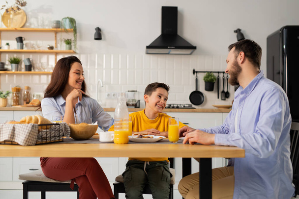 Una familia amigable desayuna en la mesa de la cocina. Un niño sonriente está sentado en la mesa con sus padres. Foto de alta calidad - Foto, imagen