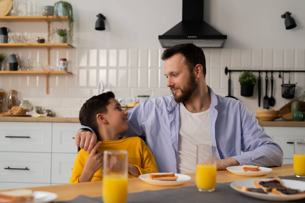 Opiekuńczy ojciec siedzi z synem przy stole kuchennym. Mały chłopiec siedzi w kuchni z ojcem jedząc kanapki. Wysokiej jakości zdjęcie - Zdjęcie, obraz