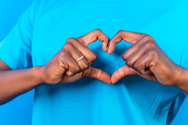 Young african american woman isolated on a blue background smiling and heart gesture, studio shoot - Photo, image