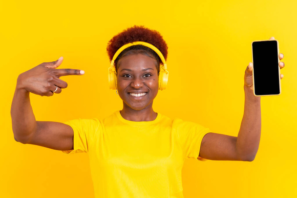 Young african american woman isolated on a yellow background with mobile pointing gesture, studio shoot - Photo, image